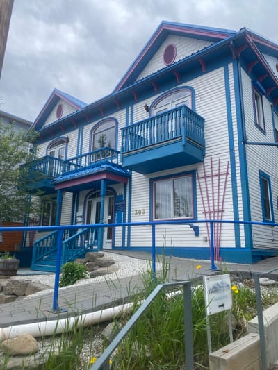 The image depicts the exterior of a two-story building in Whitehorse, Canada, with a distinctive white clapboard siding accented by bright blue and pink trim. The building features arched windows and two balconies with blue railings. A sign on the building reads "Law Offices" and "303." Another sign at the bottom right of the image says "Yukon Denture Clinic" and points with an arrow. The foreground shows a paved pathway and landscaping with rocks, grass, and some yellow flowers. The sky is overcast.