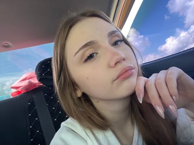 A young woman with shoulder-length brown hair, wearing a white hoodie, is photographed from the chest up, inside a vehicle. Her left hand is resting on her chin, and her right hand is visible on the right side of the frame. She has light skin, green eyes, and long, light brown eyelashes. Her lips are slightly parted. The interior of the vehicle is visible, with a black upholstered seat with a diamond pattern, a seatbelt, and a pink cloth in the background. Through the windows, a blue sky with white clouds is visible. The location is not specified as being in Марьевка, Russia.