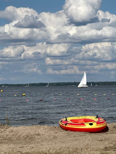 A wide-angle outdoor scene captured at a body of water in Nieporęt, Poland, likely the Zegrze Reservoir. The foreground features a sandy shoreline, with a red and yellow inflatable water tube resting on the sand at the lower right. The midground consists of dark, rippled water, marked by a visible line of small, cylindrical red and yellow buoys extending across the frame. In the distance, several sailboats are visible on the water, with one prominent white-sailed yacht positioned centrally. The background shows a dark green treeline marking the distant shore or horizon, beneath a sky dominated by large, white and grey cumulus clouds against patches of blue. No individuals are visible in the image.
