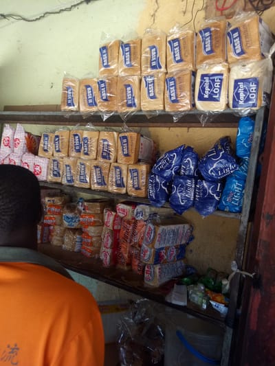 This is a medium shot, eye-level, interior photograph of a small shop or market stall. The foreground features the back and right side of a person wearing an orange t-shirt with some faded grey and black Chinese characters on the left side of the shirt. The person is facing away from the camera, towards shelves stacked with baked goods. The shelves are made of wood.
The first shelf, from top to bottom, is sparsely populated and mostly empty, except for what appear to be some packaged white pastries or bread rolls on the far left. The second shelf is densely packed with rows of packaged bread loaves. These loaves are predominantly branded as "BASE II Premium". There are also some loaves branded "Kapital LOAF" on the right side of this shelf. The third shelf contains more packaged bread and baked goods, including items labeled "Seven Star", "Sunbites", and other unidentifiable brands. There are also some plastic bags of snacks or sweets.
The background of the image consists of a textured, light yellow wall and some electrical wiring running along the top. A portion of a wooden shelf structure is visible on the right side of the image. The overall impression is of a well-stocked, if somewhat cluttered, local retail environment. The scene is likely located in Karshi, Nigeria, based on the presence of products and the general appearance of the stall.