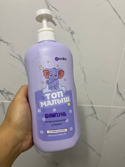 A hand is holding a light purple plastic bottle of shampoo with a white pump dispenser. The bottle has a cartoon of a purple elephant with a crescent moon and stars on it. Text on the bottle is in Russian and reads "Топ МАЛЫШ" (Top Baby), "ШАМПУНЬ" (Shampoo), "МИНДАЛЬНОЕ МОЛОЧКО И ГЛИЦЕРИН" (Almond milk and glycerin), and "от головы до пяТОЧЕК" (from head to toe). There is also text indicating the volume as "800 мл /е 27.05FL.OZ". The background appears to be a tiled wall with a marble pattern. The lighting suggests an indoor setting. No specific time of day, weather, or emotions are discernible.