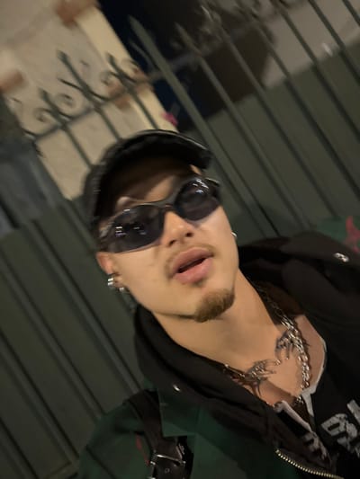 A head-and-chest level image depicts a single male individual. The subject is characterized by a light mustache and goatee, wearing dark, wrap-around sunglasses and a dark cap. Multiple silver hoop earrings are visible on his left ear. He is dressed in a dark green outer layer over a black hooded garment, featuring a prominent silver chain necklace with an intricate, stylized pendant. A black strap, consistent with a bag, is visible over his right shoulder. The subject's mouth is slightly open, and he is looking directly forward. The background is blurred, showing dark vertical elements and faint decorative metallic scrollwork, suggestive of a fence or railing. The lighting appears artificial or low-light. The scene is situated in the city of Asnières-sur-Seine, France.