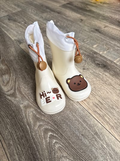 A pair of white, child-sized rubber boots with brown bear decorations and white fabric lining are placed on a wooden floor. The boots have drawstrings with wooden toggles at the top. The floor has a gray wood grain pattern. There are no people present, and the scene appears to be indoors, possibly in a home. The image does not contain any information that would allow for its location to be identified as Kondopoga, Russia.