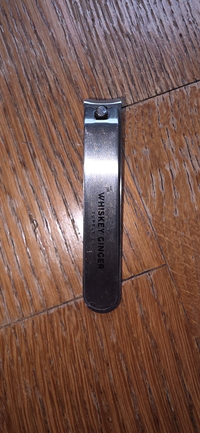 A close-up, overhead shot shows a pair of metallic nail clippers lying on a wooden surface. The nail clippers are positioned vertically in the center of the frame. The surface beneath is made of wood planks, with visible grain and texture, laid diagonally. The nail clippers have a polished metal finish and bear an inscription: "JH" and "WHISKEY GINGER SUPPLY CO.". The lighting creates subtle reflections on the metal. The context of Marum, Netherlands is not depicted in the image.