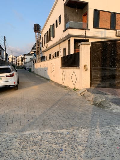 A building is visible on the left side of the shot. It has multiple windows and a dark metal gate. The facade is decorated with diamond-shaped patterns. Next to the building is a paved street with a white car parked on the side. Further down the street, more buildings can be seen. A water tank stands tall on a metal structure, and overhead power lines stretch across the sky. The sky is light blue.