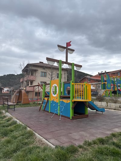 The scene depicts an empty children's playground located in Breznitsa, Bulgaria, under a uniformly overcast sky. In the mid-ground, a multi-component playground structure, designed to resemble a ship, is centrally positioned on a dark red interlocking rubber safety tile surface. This main structure features a tall mast with a red flag and two horizontal spars supporting white, semi-circular "sails." The ship-themed structure displays dominant colors of blue, yellow, green, and orange, incorporating brown wooden elements. It includes a yellow-railed upper platform, a blue slide extending to the right, a yellow panel with a white lifebuoy graphic, a brown wooden ladder, and a blue rope climbing net. To the left of the main structure, a brown wooden park bench with a dark metal frame sits beside a small, dark brown wooden tunnel. Another multi-colored playground structure, also on rubber tiles, is partially visible in the right background. The foreground consists of unkempt patches of green and dry grass, separated from the rubber surface by a low concrete curb. In the background, residential buildings with reddish-brown tiled roofs are visible, including a prominent two-story light brown house with multiple windows and bare deciduous trees in front. Treed hills line the distant horizon on the left. No individuals are present within the scene.