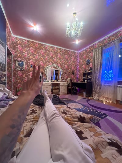 The image depicts an indoor scene, likely a bedroom, featuring floral wallpaper with pink and orange flowers. A person's legs, clad in white pants, are visible in the foreground, lying on a bed covered with a patterned blanket. The room is decorated with a chandelier and fairy lights. A vanity with a mirror, a desk with a computer, and a window with blue lighting are also present. The location is identified as Krasnoe-na-Volge, Russia, suggesting this is a residential interior within that city.