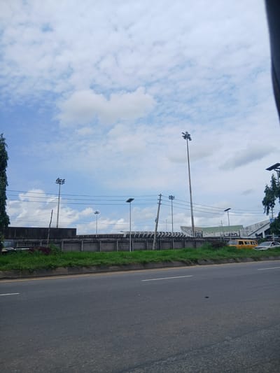 The image captures a street scene in Iganmu, Nigeria, under a cloudy sky. The foreground is dominated by a gray asphalt road with faint white markings. A concrete barrier with a grassy area behind it separates the road from a long, low-rise building complex in the background. This complex features a gray concrete wall with decorative patterns and appears to be part of a larger structure, possibly a stadium or event venue, indicated by the elevated seating visible further back. Tall, slender lampposts with multiple lights are strategically placed along the street. Several vehicles are parked or driving on the road, including a yellow minivan and a silver sedan. Lush green grass and some red-leafed plants grow along the edge of the barrier. Power lines crisscross the sky, and a tall, dark green tree stands on the far left of the frame.