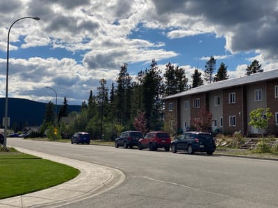 A dark blue minivan and four SUVs are parked in a paved lot next to a two-story building with brown and beige siding. The building has multiple windows and doors. Several pine trees are visible behind the building. A street light stands on the left side of the image. The sky is blue with fluffy white clouds. The scene appears to be in Whitehorse, Canada.