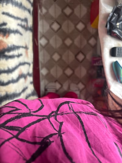 The image shows a cluttered indoor scene. A white surface with a dark plastic bag, a remote control, and a blue object sits near a floor with a geometric brown and white pattern. A tiger-striped blanket is partially visible at the bottom. In the foreground, a pink patterned cloth dominates the right side of the image. The location is not discernible as Dobrovnitsa, Bulgaria.