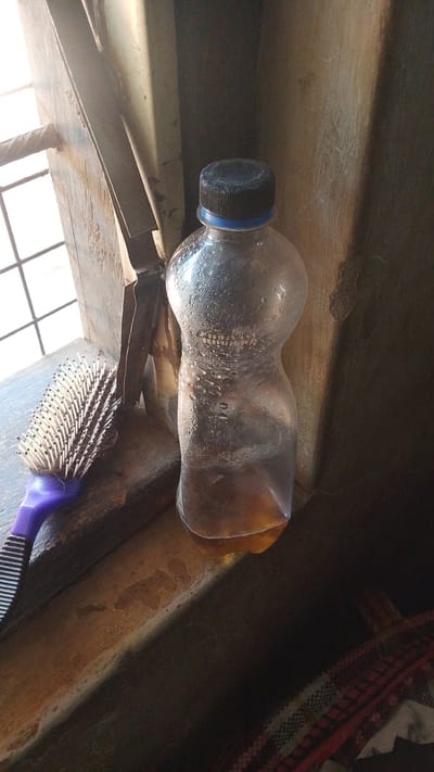 A clear plastic bottle, partially filled with a yellowish liquid, rests on a wooden surface. The bottle has a black cap with a blue ring and is covered in condensation, suggesting it's been chilled. To the left of the bottle, a purple-handled hairbrush with white bristles is also situated on the wooden surface. The background features a window with a grid of bars and a textured, darkened wall. The lighting suggests it could be daytime, with sunlight filtering through the window. The overall scene appears to be in an indoor setting, possibly a home in Donga, Nigeria. There is no discernible activity taking place; the objects are simply positioned together. Visible text on the bottle is difficult to decipher due to the condensation and image resolution but appears to be a series of numbers and letters.