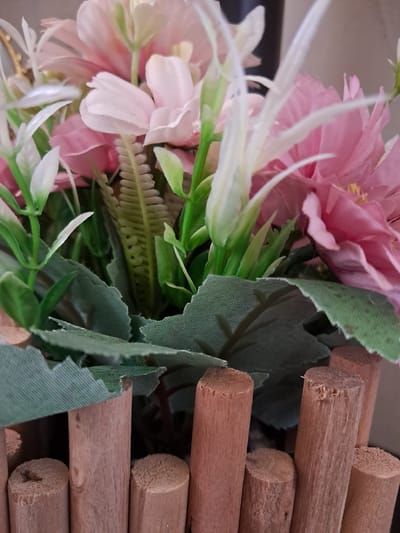 This close-up image captures a static arrangement of decorative items, likely situated indoors. On the left side, a vibrant display of artificial flowers dominates, featuring soft pink and white petals made from fabric or synthetic materials. Delicate white, elongated elements, possibly stamens or pistils, emerge from the blooms. Interspersed among the flowers are various artificial green leaves; some are smooth and broad, while others exhibit a finely ridged, fern-like texture, suggesting a mix of synthetic foliage.
To the right of the frame, contrasting with the soft textures of the flowers, a series of natural wooden dowels or rods are stacked horizontally. These cylindrical pieces of wood are light brown, showing natural grain patterns and slightly coarse, cut ends. The composition highlights the juxtaposition of the delicate, artificial floral elements with the rougher, natural wood.
There is no visible activity taking place, nor is there any discernible text within the image. The lighting suggests an indoor setting, and no specific time of day or weather conditions can be inferred. The elements presented are generic decorative items, and the image itself offers no specific geographical markers to indicate its location in Gavar, Armenia, beyond the provided context.