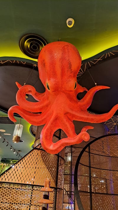 An overhead shot captures a vibrant, oversized orange octopus sculpture suspended from a ceiling. The sculpture is highly detailed, with textured skin and distinct eye sockets, and its numerous tentacles spread out in various poses. The octopus is positioned against a backdrop of dark material, some of which has a geometric pattern in gold.
To the left of the octopus, a portion of a large, dark green surface is visible. It has a curved edge and a bright yellow accent line. A small, circular white fixture with a black square symbol is mounted on this green surface. Below this, a circular ventilation grate with concentric rings is also visible, emitting a faint yellow glow.
The background is dominated by intricate metal latticework, casting dappled light and creating a sense of depth. The latticework is made of gold-colored metal and features a variety of patterns, including honeycomb and scalloped designs. Some areas of the latticework are illuminated with a purple and orange light, suggesting artificial lighting within the space. A few clear glass pendant lights with brass accents hang among the latticework.
The overall impression is of an indoor space, likely a restaurant or entertainment venue, with a playful and artistic theme. The lighting is subdued, contributing to a sophisticated atmosphere. The scene appears to be well-maintained and designed for visual appeal. There is no visible text in the image.