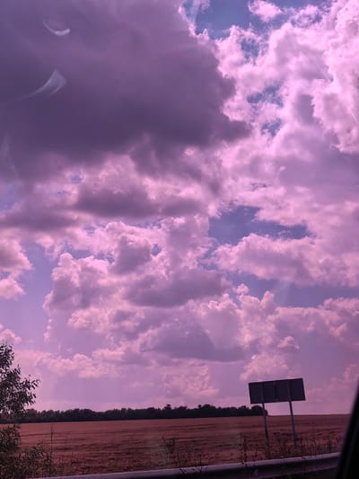 The image shows a rural landscape with a field in the foreground and a treeline in the background. The sky is filled with dramatic, pink-hued clouds. There is a dark, rectangular signpost with two blank panels in the right side of the image. The overall color tone of the image is heavily influenced by a purplish-pink cast, suggesting either a filter or unusual lighting conditions. A metallic guardrail runs along the bottom of the frame. The location is identified as Churtan, Russia, but there are no specific urban features visible, indicating this is a rural area within or near the settlement.