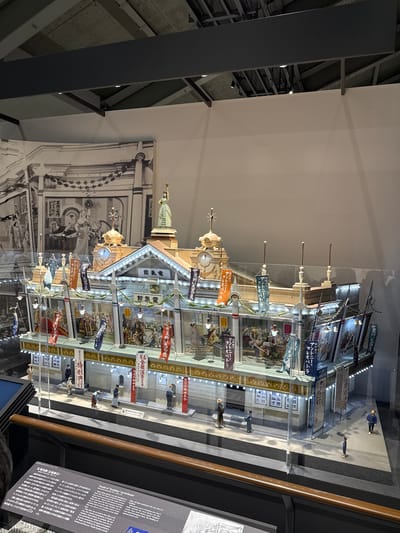 The image depicts a detailed diorama or miniature model within a glass display case. The model appears to be of a building or structure adorned with numerous banners featuring Japanese text, portraits, and illustrations. Small figurines are positioned within and around the model, representing people in period attire.
The diorama is part of an exhibition, with a large monochrome photograph in the background that complements the model. Text panels providing information about the display and its historical context are visible on the right side of the image. The setting is indoors, likely a museum or exhibition hall, as indicated by the architectural details of the ceiling and support structures.
Based on the OCR text provided, the exhibit refers to the history of "Denkikan" movie theaters in Japan, potentially located in the Sumida Ward, Tokyo. The diorama appears to be of such a theater.
