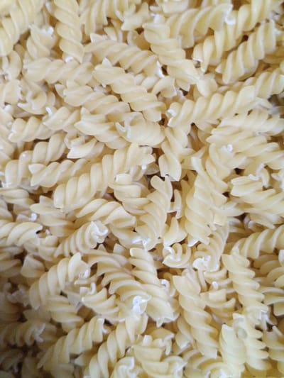 The image is a close-up, overhead view of a large pile of cooked fusilli pasta. The pasta is a pale yellow color and appears to be slightly glossy, suggesting it has been recently cooked or tossed with oil. The spiral shapes of the fusilli are clearly visible, with many pieces overlapping each other. There are no people or other objects present in the image, and the background is entirely filled with pasta. The scene is indoors and well-lit.
The location provided is the city of Martuni, Armenia. Without further information or visual cues within the image itself, it is impossible to pinpoint the exact location within Martuni. It could be a kitchen in a home, a restaurant, or a food production facility.