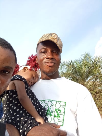 A close-up outdoor selfie captures three individuals under a clear blue sky, likely in Omicha Umu Awada, Nigeria. In the foreground, a man with dark skin, wearing a camouflage baseball cap and a white t-shirt, looks directly at the camera with a subtle smile. He is holding a young child, who appears to be a girl, fast asleep with her head resting on his chest. The child has red braided hair with a white headband and is dressed in a black and white polka dot outfit. To the far left, a portion of another person's face, including an eye and forehead, is visible, suggesting they are very close to the camera, possibly taking the photo.
The setting is bright and sunny, indicating daytime. In the background on the right, a vibrant green palm tree and other distant foliage are visible. The man's white t-shirt features a prominent green embroidered logo with a geometric, chain-link-like pattern, enclosing what appear to be Greek letters "A" (Alpha) and "Σ" (Sigma). The overall mood is serene and casual.