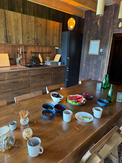 The image depicts a rustic, wooden-paneled kitchen or dining area, likely situated in a rural or cabin-like setting. A long, wooden table dominates the foreground, set with various food and drink items. On the table, there are several white ceramic mugs, some with spoons, a glass jar containing dried herbs, a small jar of honey, a decorative glass holder filled with breadsticks, and several blue bowls containing dried fruits and nuts. A plate of sliced watermelon is centrally placed, with a small dish of raspberries nearby. To the right, three green glass bottles of sparkling water stand alongside two textured glass tumblers.

In the background, wooden cabinetry lines the wall, with a black refrigerator to the left. A sink and faucet are visible above the counter. A framed "VITAMIN CHART" hangs on the wooden-paneled wall. The lighting appears to be warm, with visible overhead fixtures and a glowing decorative lamp to the left of the refrigerator. Wooden chairs are partially visible around the table. The overall impression is one of a cozy, communal space prepared for a meal or snack. The mention of Dobrzyń, Poland, suggests a possible location, but there are no specific visual cues in the image to confirm this geographical detail.
