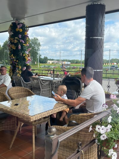 This is an outdoor shot taken at a cafe in the village of Akhlebaikha (Kovriginskii S/S), Russia. The scene is set on a covered patio area. In the foreground, a glass-top wicker table is set with a phone and a remote. A man in a grey t-shirt is seated in a wicker chair, holding a phone and looking at it. Next to him is a baby in a stroller. In the background, several other people are seated at tables. There is a decorative column covered in flowers to the left of the frame, and another column wrapped in a black mesh to the right. The area overlooks a road with parked cars and greenery, under a cloudy sky.