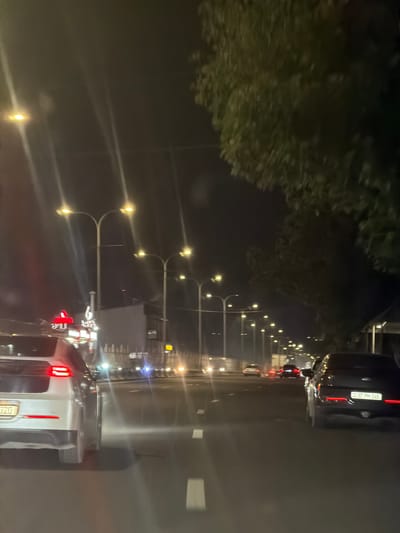 The image depicts a night-time scene on a multi-lane road in Parakar, Armenia. The perspective is from inside a vehicle, looking forward and slightly to the right. The road is illuminated by numerous streetlights, creating streaks of light due to the long exposure. There are several cars on the road, some of which are visible in the foreground and middle ground, all moving in the same direction. On the left side of the road, there are buildings and illuminated signs, including a red sign with white lettering and a white sign with dark lettering. Trees with dense foliage are visible on the right side of the road, their leaves illuminated by the streetlights. The road surface is asphalt, and white lane markings are visible. The overall impression is a typical urban nighttime driving scene with moderate traffic.
