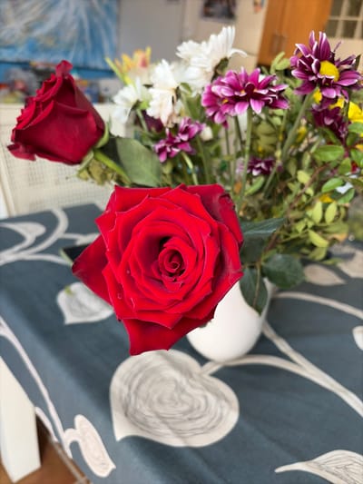 This image depicts a floral arrangement situated indoors on a table in the city of Benahavís, Spain. In the foreground, a prominent red rose, exhibiting several water droplets on its petals, is centered slightly to the left. Immediately behind it, a white, opaque, bulbous vase holds a mixed bouquet of flowers. This bouquet contains at least two additional red roses, multiple white daisy-like flowers, and several purple/magenta chrysanthemum-like flowers with yellow centers, interspersed with green foliage. The table is draped with a dark blue/grey tablecloth patterned with repeating light grey/white stylized heart-shaped forms and swirling organic lines. The background, largely out of focus, reveals a light-colored chair back on the left, an abstract blue and white artwork or poster on a wall, and blurred outlines of wooden furniture or cabinetry on the right. No individuals are present in the scene, and no discernible actions or interactions are occurring.