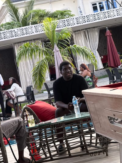The photograph captures a social gathering at a poolside venue in Rubuchi, Nigeria. In the foreground, a young man with dark skin and short black hair is seated, wearing a black t-shirt and grey shorts. He holds a plastic bottle of water. In front of him is a low, rattan coffee table with a glass top, on which rests a glass jar containing a beverage with mint leaves and a straw. A large red cushion is visible on a rattan chair to his left. Behind him, a woman with long, auburn hair is seated at another table, wearing a strapless green and white patterned dress. She has gold hoop earrings. Further in the background, other individuals are present, and a maroon umbrella is set up. The environment suggests an outdoor lounge area with potted plants and curtains. The architecture in the background appears to be part of a building with white decorative elements and windows.