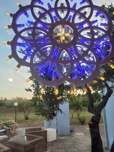 The image, taken in Sogliano Cavour, Italy, depicts an outdoor evening scene focused on a decorative light fixture. The primary subject is a large, elaborate light ornament, possibly a chandelier or garden decoration, constructed from interlocking white circular or wheel-like segments. It is adorned with numerous small LED bulbs; many are a vibrant blue, while others emit a warm, golden-yellow light. Some clear bulbs are also visible.

Behind and around the light fixture, the scene opens to an outdoor setting at dusk. A tree with dark, gnarled branches and green leaves is prominent in the background, creating a natural backdrop. Beyond the tree, an expansive field or garden area is visible, with grass and more distant trees under a twilight sky. A large, bright, circular orb, likely the moon, is visible in the upper-right portion of the sky.

To the right side of the frame, there is a paved patio area furnished with several pieces of outdoor wicker furniture, suggesting a seating or lounge space. A white, square-shaped outdoor light or planter is also present on the patio. Further into the distance, a lamppost with a spherical light illuminates a patch of white flowers. The overall ambiance is tranquil and inviting, characteristic of an evening in an outdoor leisure area. There is no visible text in the image.