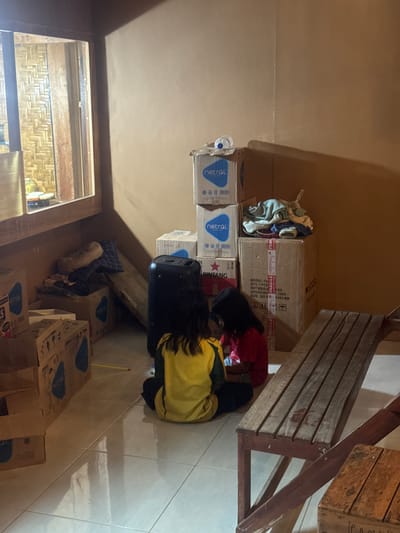 In the city of Kertasari, Indonesia, an indoor scene is depicted, likely a storage area or room. Two young girls, one wearing a yellow and green shirt and the other a red shirt, are seated on the floor amongst stacks of cardboard boxes. The boxes, some bearing the "netral" logo and "BINTANG" branding, suggest potential goods for storage or transport. A large black speaker or electronic device stands to the left of the girls. A wooden bench is positioned in the foreground, to the right, with an open wooden crate beside it. The walls are a warm, earthy tone, and the floor is tiled. Natural light filters in from a window on the left, revealing a woven bamboo texture behind a wooden frame. The overall impression is one of a temporary storage space or a home environment with unpacked or stored items.