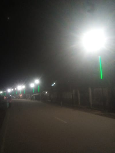 The image depicts a street scene at night in Tundun Wada West, Nigeria. A bright street lamp dominates the right side of the frame, casting a harsh glare and creating lens flare. To the left of the lamp, a series of streetlights are visible, interspersed with green vertical lights. The street itself is paved and appears to be a road, with indistinct figures and vehicles present, suggesting activity. The overall impression is one of a well-lit urban thoroughfare at night.
