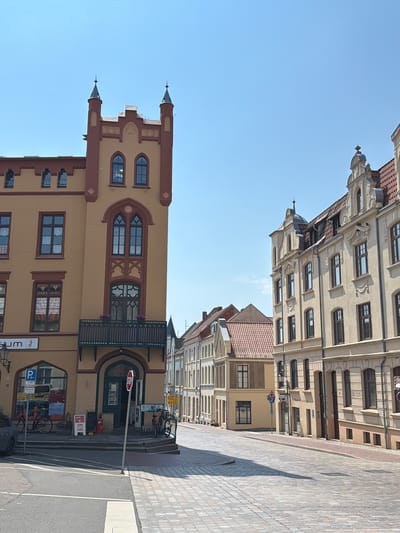The image shows a street scene in the Hanseatic city of Wismar, Germany, specifically the intersection of Bergstraße and Kirchplatz. On the left is a prominent yellow building with red trim, featuring gothic architectural elements like pointed arches and towers. This building appears to be a historical structure, possibly housing shops or a museum on the ground floor, indicated by signage like "um". A blue parking sign is visible, along with bicycles and a car. A red and white stop sign stands on the sidewalk.
To the right of the yellow building, the street, paved with cobblestones, curves uphill. Along this street are several other historic buildings, characterized by their light-colored facades, red tiled roofs, and detailed architectural ornamentation, typical of northern German brick Gothic and Renaissance styles. These buildings are multistory, with numerous windows. The sky is clear and blue, suggesting a sunny day. No people are directly visible engaging in any actions, though parked bicycles and a car indicate present activity. The scene depicts a quiet urban environment focused on historic architecture.