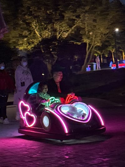The image depicts a group of people posing next to a neon-lit amusement park ride. The ride is shaped like a car with neon outlines. The people are standing or sitting near the ride, some looking at the camera. The setting appears to be at night, possibly at an amusement park. There are no details to determine the location in the city of Jinzhou, China.