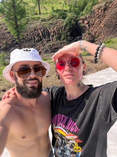 In Talnakh, Russia, a man and a woman are taking a selfie outdoors. The man, positioned on the left, wears a white bucket hat with an Adidas logo and aviator sunglasses. He has a beard and is shirtless. The woman, on the right, wears round red sunglasses, a nose ring, and has plugs in her ears. She has tattoos on her arm and neck. She is wearing a dark t-shirt with a graphic print featuring mushrooms and text. The couple is embracing, with the woman's hand resting on the man's shoulder and his arm around her. They are in a natural setting with a rocky, tree-covered hillside in the background and a patch of white ground, possibly snow or ice, in the foreground.