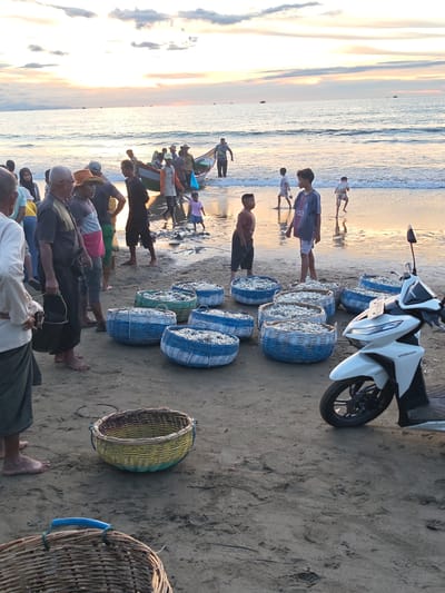 The image depicts a beach scene in Uleekareung, Indonesia, during sunset. Numerous people, including adults and children, are present on the sandy shore and in the shallow water. Several fishing boats are visible in the ocean, with some individuals interacting with them. Large woven baskets, filled with fish, are arranged on the sand. A white scooter is parked on the beach. The sky displays a gradient of colors from the setting sun, with clouds present. The environment suggests a fishing community actively engaged in its livelihood.