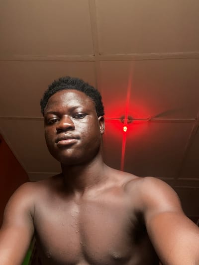 A shirtless, muscular, dark-skinned male with short, curly black hair is positioned in front of a wall with vertical panels. The location is difficult to definitively determine, but given the prompt, the location would hypothetically be in the city of Mosafejaw, Nigeria. There is a red light fixture on the wall, casting a red glow. The subject appears to be glistening with sweat or moisture. The photograph is taken at an angle.
