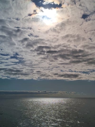 The image depicts a body of water, likely a lake or sea, under a sky filled with layered clouds. The sun is visible through the clouds, with a slight rainbow effect around it. The water reflects the sunlight, creating a shimmering effect. In the distance, a faint outline of land can be seen on the horizon. There are no people visible in the image. The exact location cannot be definitively determined as being in Ozernoe Ust'e, Russia, as the image contains no identifying markers specific to that location.