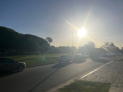The image depicts an outdoor urban scene in the city of Partido de Quilmes, Argentina, during daytime, characterized by a low sun angle indicating either morning or late afternoon. A clear, light blue sky dominates the upper portion, transitioning to intense brightness around a prominent sunburst in the upper right quadrant, which casts significant lens flare.
The scene features an asphalt-paved road running diagonally from the lower left foreground into the mid-background. On the far left, a dark-colored sedan or hatchback is partially visible, either parked or in slow motion, facing away from the viewer. Its side profile and front wheel are discernible.
To the right of the road, a narrow grassy median strip is separated by a concrete curb. Beyond this, a section of cobblestone pavement is visible in the lower right foreground. Further along the road on the right, two compact cars are visible in the mid-distance; one is light-colored, the other dark-colored, both appearing to head away from the viewer. Adjacent to these vehicles, a dark, rectangular utility structure is present. A line of several light-colored vehicles, possibly vans, is parked or moving further down the road in the background on the right, partially obscured by distance and sun glare.
To the left of the road and extending into the background is a large expanse of manicured green grass, indicative of a park or public green space. This area is bordered by a dense line of mature trees with dark foliage, silhouetted against the brighter sky. Faint, dark human-like figures are discernible within the grassy area near the trees. Two streetlights with dual luminaires are silhouetted against the bright sun in the mid-background. All visible vehicles and stationary objects cast long, distinct shadows towards the lower-left quadrant of the frame.