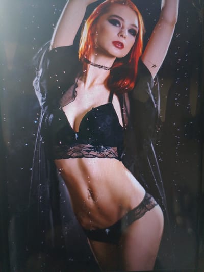 The image displays a full-body shot of a woman with vibrant red hair, styled in a way that emphasizes her striking facial features. She is wearing black lingerie, consisting of a lacy bra and matching underwear, along with a sheer black robe. Her arms are raised, creating a dynamic pose that highlights her slender physique, including her toned abdomen and a navel piercing. The background is dark and appears to be a studio setting, with a soft focus that draws attention to the subject. Small, scattered points of light create a magical or ethereal effect, resembling glitter or falling stars. The woman's expression is alluring, with bold makeup including dark eyeshadow and red lipstick. The overall mood is sensual and mysterious, with a hint of a dramatic flair due to the lighting and pose. There is no visible text in the image. The location context of Dobra Krynytsia, Ukraine, does not provide any specific visual cues within the image itself.
