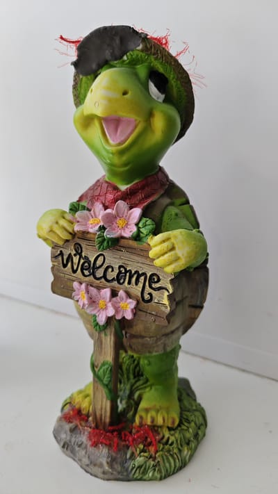 A green, cartoonish turtle figurine stands upright, holding a wooden sign that reads "Welcome" in black script. The turtle is wearing a straw hat with a black band and red frayed material sticking out, a reddish scarf, and a brown vest. Pink flowers with yellow centers are attached to the sign and around the turtle's neck. The figurine is situated on a textured green base representing grass and a rock, with red string-like material scattered on the grass and rock. The background is a plain white surface. The figurine is a decorative item, possibly for a garden or indoor display. There is no discernible information regarding its location in Shemordan, Russia.