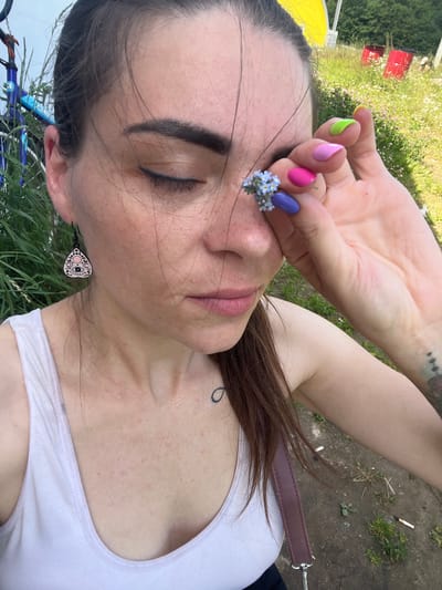 A woman, likely in her late 20s to early 30s, with brown hair, closes her eyes as she holds a small cluster of blue forget-me-not flowers to her left eye. She wears a white tank top and a dark earring with a white and pink moon and eye design. Her fingernails are painted in various bright colors, including neon green, pink, and purple. A tattoo is visible on her upper chest and on her left arm. The background suggests an outdoor, possibly rural, setting with tall green grass, a dirt path, and some red barrels and a yellow structure in the distance. A bicycle is partially visible on the left side of the frame. The lighting indicates it is daytime.