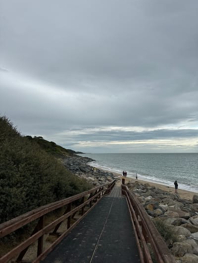 This image depicts a wooden walkway leading down to a beach in Courtown, Ireland. The walkway has a black, textured surface and is flanked by wooden railings. On the right side of the walkway, there is a rocky embankment. The beach itself is sandy and appears to be partially covered with rocks. The sea is calm with gentle waves, and the sky is overcast with gray clouds. Several people are visible on the beach. In the background, there is a grassy, vegetated hill.