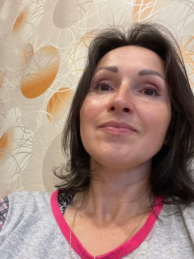 The image is a close-up, low-angle selfie of a woman with dark hair and brown eyes. She is wearing a grey t-shirt with pink trim and a thin gold necklace. The background is wallpaper with an abstract pattern of circles and lines in shades of white, grey, and orange. The lighting suggests an indoor setting. The woman's face is slightly turned upwards and to the right, with her gaze directed off-camera. There are no identifiable landmarks or clues within the image to pinpoint a specific location in Kumyon, Russia.