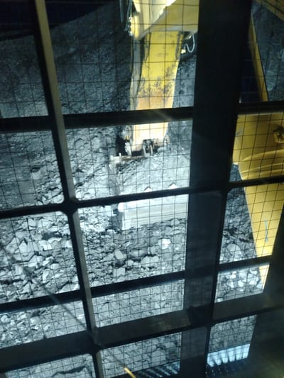 The image shows a view from above, through a metal grate, of a mining operation. The ground is covered with rubble and rocks, and a yellow piece of heavy machinery, likely a loader or excavator, is visible. The machine appears to be in operation, as there are indications of disturbed earth and debris. The scene is dimly lit, suggesting it is either night or underground. The overall impression is one of an active industrial site. The specific location in Naminga, Russia, is not depicted in the image itself.