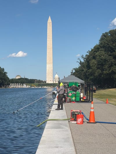 The image depicts the Washington Monument and the Reflecting Pool in Washington, D.C. On the right side of the frame, near the edge of the Reflecting Pool, a man in a gray work uniform and a bright yellow hard hat is operating equipment. He is standing next to a red gas-powered pump and an orange Home Depot bucket. Two large green containers are positioned behind him, under a grey canopy. A fence encloses some of the equipment. An orange traffic cone is visible next to the pump. A blue hose runs from the pump and disappears out of frame to the right. A green hose extends from the water of the Reflecting Pool towards the pump. Another person in dark clothing is partially visible in the background near the water. In the distance, across the Reflecting Pool, the Lincoln Memorial, the World War II Memorial, and the U.S. Capitol Building are visible. The sky is blue with a few scattered white clouds. Trees and grassy areas form the background on the right side of the image.