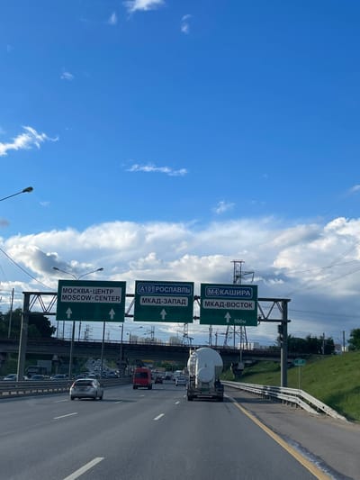 The image depicts a section of highway in Novodrozhzhino, Russia, showing several green road signs indicating directions to various destinations. The signs, written in both Russian and English, point to "Moscow-Center," "A101 Roslavl," "MKAD-Zapad (West)," "M4 Kashira," and "MKAD-Vostok (East)." A distance of "500m" is noted on one sign.
The highway appears busy with cars and trucks moving in both directions. Power lines are visible above the road. The sky is predominantly blue with white clouds. A green grassy area runs alongside the highway.