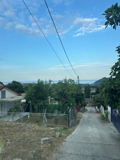 The image depicts a narrow, paved road winding downhill through a residential area. On the left, a white house with a red roof is visible, partially obscured by a chain-link fence and dry grass. Further down the road, several trees with lush green foliage line both sides, casting shadows on the asphalt. The scene opens up to a vista of the sea in the distance, with a hazy horizon and a clear blue sky dotted with white clouds. A prominent power line stretches across the upper portion of the frame. The overall setting appears to be a quiet, rural or semi-urban environment.