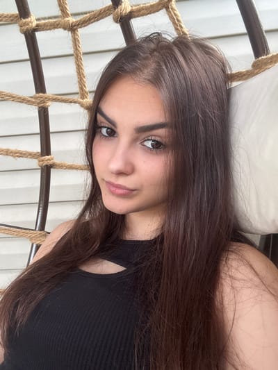The image shows a young woman with long, dark brown hair lying down in a hanging chair. She is wearing a black tank top. Her head is tilted, and she is looking directly at the camera with a soft expression. The background consists of a white wall with vertical siding and some rope details from the chair. The lighting suggests it is daytime, possibly outdoors, given the natural light. The overall scene appears casual and relaxed. There is no visible text in the image.