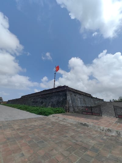 An outdoor, eye-level wide shot depicts a large, weathered stone fortress wall, or rampart, under a partly cloudy blue sky in Phú Xuân Ward, Vietnam. The wall, dark gray and appearing constructed of blocks, extends horizontally across the middle of the frame from left to right, turning at a corner on the right. A prominent multi-segmented flagpole is mounted atop the wall to the left of the corner, flying a red Vietnamese flag with a central yellow five-pointed star.

In the foreground, the ground is paved with reddish-brown square tiles. A raised, light-colored curb separates this area from a darker, possibly gray, paved path that runs diagonally towards the upper left, parallel to the wall. A strip of green ground cover, possibly low shrubs or vines with scattered yellow flowers, borders the base of the fortress wall on the left side of the frame. To the right, a dark metal fence with decorative scrollwork separates the tiled foreground from an upward-sloping, dark-colored stone or paved ramp leading to the top of the fortress. No people, discernible actions, or interactions are visible in the scene. The overall lighting suggests a bright, sunny day.
