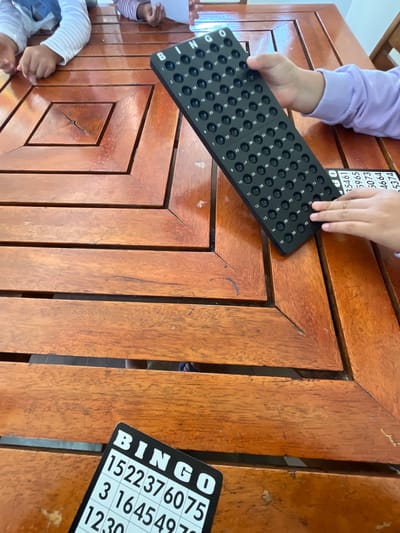 On a wooden table, children are playing Bingo. One child holds a black Bingo board with numbered holes. A black Bingo card with numbers is on the right side of the table. A partially visible Bingo card is at the top left. This activity is taking place on a wooden table in an unknown location in the city of Pacarán, Peru.
