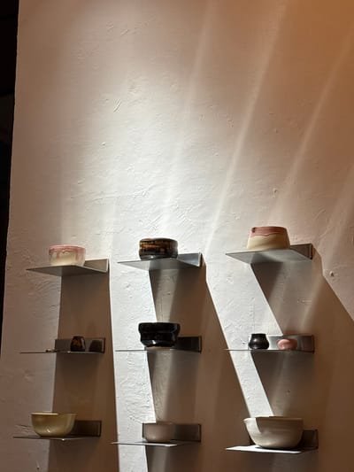 Ten ceramic vessels are displayed on nine flat, rectangular metal shelves mounted against a textured, light-colored (off-white or cream) wall. The wall surface exhibits subtle textural variations, consistent with plaster or stucco. Overhead or angled artificial lighting illuminates the display, casting prominent diagonal shadows from the shelves and objects downwards and to the right across the wall.

The shelves are arranged in three vertical columns, each containing three shelves.

On the top row, from left to right: a cream-colored bowl with pink glaze dripping over its rim; a dark, rounded bowl with a complex, multi-toned, earthy glaze concentrated on its rim; and a cream-colored bowl with a smooth, soft pink band encircling its upper portion.

The middle row displays, from left to right: a small, dark vessel with an irregular, organic shape, possibly in a dark brown or reddish hue; a dark, black-glazed bowl with distinct vertical fluting or corrugated texture; and two smaller items—a dark, reflective, cup-like vessel and a smooth, ovular, light pink object.

The bottom row features, from left to right: a wider, shallow bowl with a uniform pale yellow or beige glaze; a small, shallow bowl with a uniform pale pink or cream glaze; and a larger, deep bowl with a uniform pale beige or off-white glaze.

No human figures, actions, or interactions are depicted within the scene. The precise location within the city of Guadalajara, Mexico, is not discernible from the visual evidence provided.