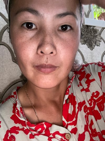 The image is a close-up, eye-level, indoor selfie of a woman in Sary-Kamysh, Kyrgyzstan. The woman is looking directly at the camera. She has dark hair pulled back and is wearing a light-colored shirt with a red floral pattern and a gold chain necklace. Her skin has freckles. The background consists of a patterned wallpaper and a glimpse of greenery through a window. The lighting suggests a natural light source.